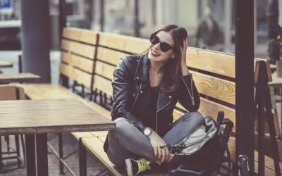 Dziewczyna w skórzanej kurtce i okularach przeciwsłonecznych, z uśmiechem na twarzy, siedzi na ławce w kawiarni. Jej styl jest zdecydowanie rockowy.