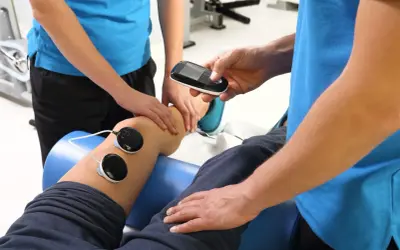 Fizjoterapeuta wykonuje zabieg fonoforezy na kolanie pacjenta, używając aparatu i elektrod.