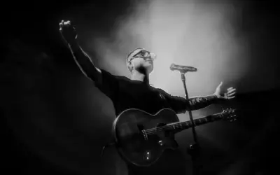 Artysta z gitarą na scenie, w okularach, z podniesionymi rękami.