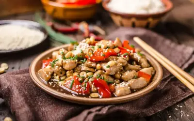 Pyszny kurczak po turecku z orzeszkami ziemnymi i papryką, posypany sezamem i szczypiorkiem. Obok miseczki z ryżem i pałeczki.