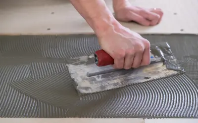 A hand uses a notched trowel to spread thinset for cement board, creating a textured surface for tile adhesion.
