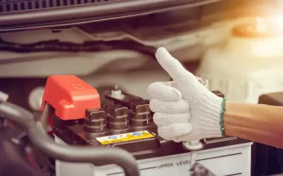 Main d'une personne portant un gant blanc fait un pouce levé près d'une batterie de voiture. Une astuce pour le démarrage à froid diesel.