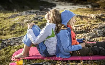 Två personer, en vuxen och ett barn, sitter rygg mot rygg på en liggunderlag i naturen. En härlig stund att vandra med barn.
