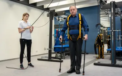 Starszy mężczyzna w specjalistycznej uprzęży i z kulami ćwiczy pod okiem fizjoterapeutki. To kluczowy etap rehabilitacji neurologicznej.
