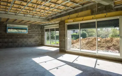 Wnętrze w budowie z betonową podłogą i ścianami. Widoczna konstrukcja sufitu i okna z widokiem na plac budowy. Powstaje nowa ściana działowa.