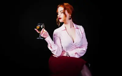 Donna con capelli rossi tiene un calice di vino da meditazione, con un'espressione pensierosa. Indossa una camicetta rosa e una gonna bordeaux.