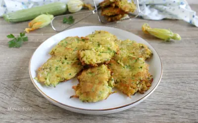 Buñuelos de calabacín dorados y crujientes en un plato blanco, con calabacines frescos y flores en el fondo.