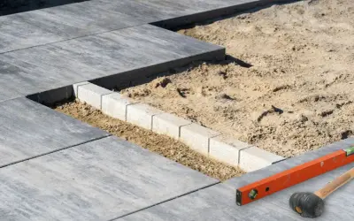 Budowa tarasu z płyt betonowych w toku. Widać układane płyty, podsypkę piaskową i obrzeże. Poziomica i gumowy młotek czekają na dalsze prace.