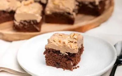 En bit chokladtårta med krämig frosting och nötter. Flera bitar serveras på ett träfat i bakgrunden.