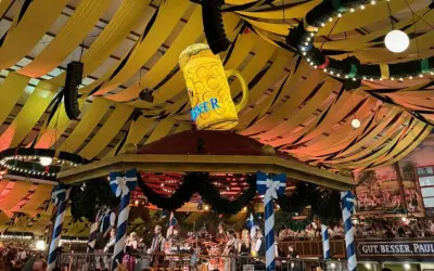 Wielki, świecący kufel piwa wisi nad sceną z zespołem. Dekoracje w kolorach biało-niebieskim i żółtym tworzą atmosferę Oktoberfest 2025.