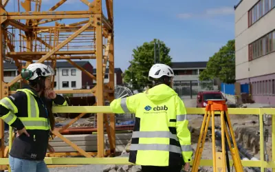 Två glada medarbetare i varselkläder och hjälmar på en byggarbetsplats. En stor kran syns i bakgrunden.