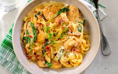 En skål med krämig pasta med soltorkade tomater, kyckling och spenat, toppad med riven parmesan och färska örter.