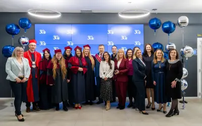 Grupa absolwentów i wykładowców świętuje 30-lecie swojej alma mater. Wszyscy ubrani są w togi i birety, a w tle wiszą balony.