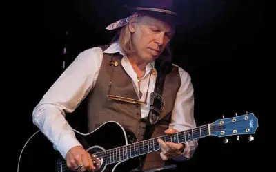 Elliott Murphy Zaragoza, con sombrero y chaleco, toca la guitarra acústica en un concierto.