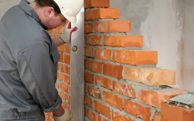 Murarz w kasku i rękawicach używa poziomicy do sprawdzania równości różnych rodzajów cegieł podczas budowy ściany.