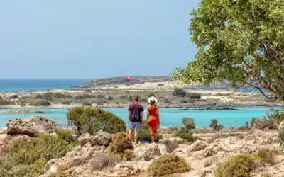 Ett par njuter av utsikten över Elafonissi, Kreta, med dess turkosa vatten och steniga landskap. En röd fallskärm syns i fjärran.