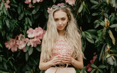 Młoda kobieta o blond włosach i słowiańskiej urodzie, w kwiecistej sukience, stoi na tle bujnej zieleni i różowych kwiatów.