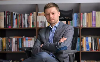 Mężczyzna w marynarce siedzi na krześle przed regałami pełnymi książek. Zastanawiasz się, ile książek ma Remigiusz Mróz?