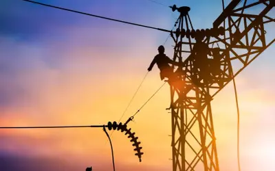 Sylwetka pracownika na tle zachodzącego słońca, naprawiającego linię energetyczną. Widać izolatory i konstrukcję wieży, a także potencjalny łuk elektryczny.