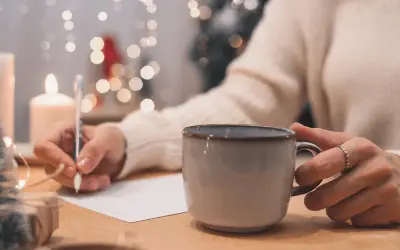 Osoba pisze podpis pod życzeniami urodzinowymi, trzymając kubek z gorącym napojem. W tle migoczą światełka choinkowe.
