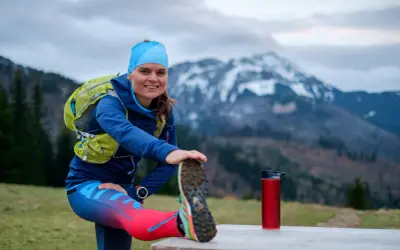 Uśmiechnięta biegaczka przygotowuje się do treningu ultramaraton 50 km, rozciągając nogę na tle gór.