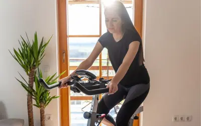 Młoda kobieta ćwiczy na rowerku stacjonarnym, zastanawiając się, czy rowerek stacjonarny odchudza. Słońce wpada przez okno.