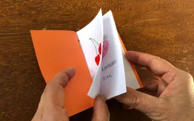 Mani che sfogliano un piccolo libro fatto a mano, con disegni di fragole e ciliegie. Un'idea per imparare come fare un libro a mano.