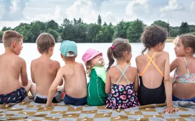 Dzieci siedzą na kocu nad jeziorem, ciesząc się fajnymi wczasami. Jedno z nich ma lody na twarzy.