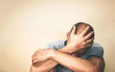Mężczyzna zakrywa twarz dłońmi, siedząc przy jasnej ścianie.