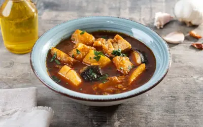 Un tazón de patatas con bacalao en salsa, decorado con perejil. Al lado, una botella de aceite de oliva y ajos.