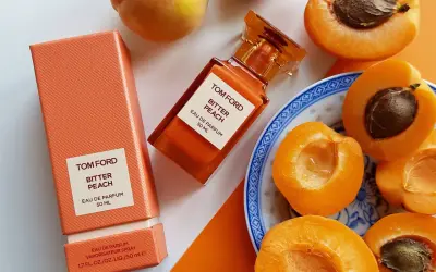 Pomarańczowe opakowanie i buteleczka perfum Tom Ford Bitter Peach otoczone soczystymi brzoskwiniami.