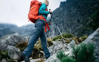 Kobieta z plecakiem wyrusza na wypad w góry, wspierając się kijkami trekkingowymi po kamienistym szlaku.