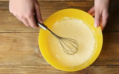 Mains préparant la pâte à crêpes dans un bol jaune. La pâte est prête pour son temps de repos.