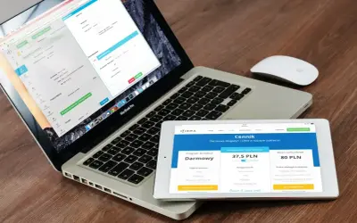 Programista .NET pracuje nad aplikacją na laptopie i tablecie. Na ekranach widoczne są formularze i cennik usług.