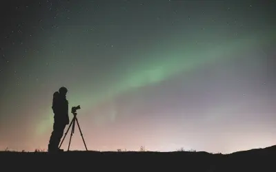 Sylwetka fotografa z aparatem na statywie pod rozgwieżdżonym niebem i zorzą polarną. Efekt bokeh gwiazd dodaje magii.
