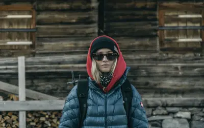Kobieta w czerwonej bluzie z kapturem i czarnej czapce, w okularach przeciwsłonecznych, z plecakiem, przed drewnianą chatą.