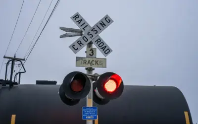 Przejazd kolejowo-drogowy z czerwonym światłem. Znak "Railroad Crossing" i informacja o 3 torach. W tle czarny wagon kolejowy.