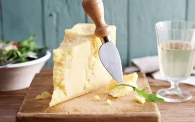 Ein Stück Parmesan, perfekt für vegetarische Gerichte, wird mit einem Käsemesser angeschnitten. Daneben ein Glas Weißwein und ein Salat.