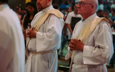 Diakoni i księża w białych szatach, z modlitewnie złożonymi dłońmi. Różnica między diakonem stałym a księdzem, ich obowiązki i przywileje.
