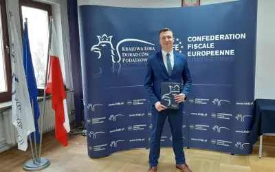 Mężczyzna w garniturze stoi przed niebieskim banerem z logo Krajowej Izby Doradców Podatkowych.