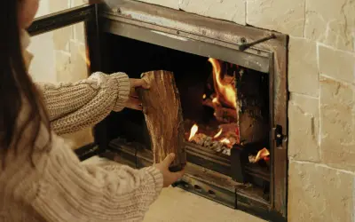 Une personne en pull ajoute une bûCHE dans un poêle à bois qui fume, porte ouverte. La chaleur des flammes réchauffe la pièce.