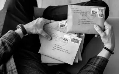 Ręce trzymające stos brązowych kopert, każda z adresem i znaczkiem pocztowym. Wygląda jak przygotowanie do wysyłki listów poleconych ekonomicznych.