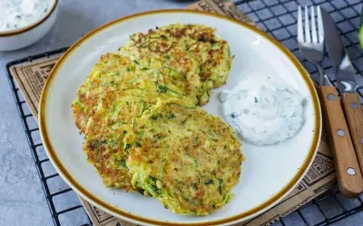 Drei goldbraune Zucchini-Pfannkuchen liegen auf einem Teller, daneben ein Klecks Kräuterquark. Ein leckeres Gericht!