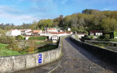 Antiguo puente de piedra con señal de peregrinación. Al fondo, un pueblo con casas de tejados rojos y vegetación exuberante. ¡Ponte Maceira que ver!