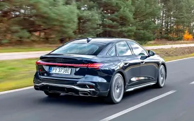Ciemny Audi A4 B10 w wersji sedan pędzi po drodze, otoczony jesiennym lasem.
