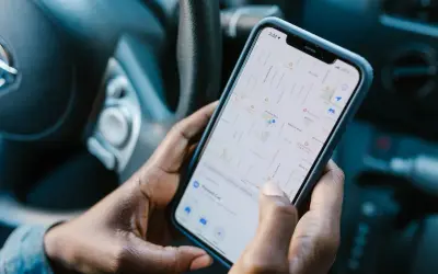 Dłonie trzymają telefon z mapą, pokazującą aplikację do udostępniania lokalizacji. Widać fragment kierownicy samochodu.