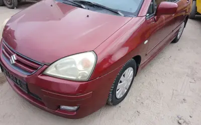 Czerwony Suzuki Liana stoi na piaszczystym podłożu. Widoczny przód auta z reflektorem i grillem.