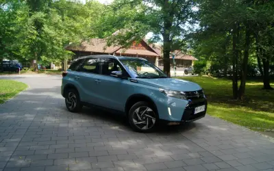 Jasnoniebieski Suzuki Vitara Hybrid zaparkowany na wybrukowanym podjeździe, otoczony zielenią i drewnianym budynkiem.