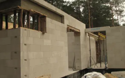 Budowa domu z bloczków betonowych. Widoczna ściana nośna z otworami okiennymi i drzwiami, podparta drewnianymi belkami.