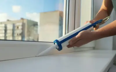Ręce trzymają pistolet z uszczelniaczem, aplikując go na biały parapet okna.